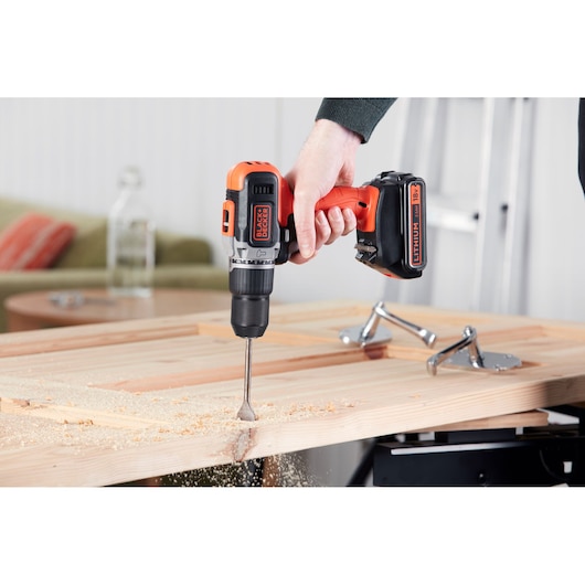 BLACK+DECKER BCD003ME2K cordless drill in use, drilling into a wooden plank indoors.