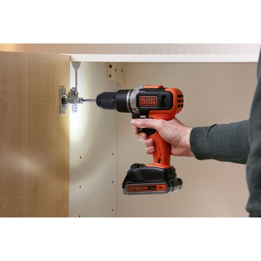 BLACK+DECKER BCD003ME2K cordless drill in use installing a cabinet hinge, orange and black design visible.