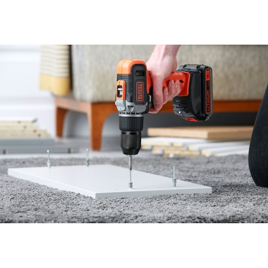 BLACK+DECKER BCD003ME2K cordless drill in use, drilling screws into a white board on a carpeted floor.