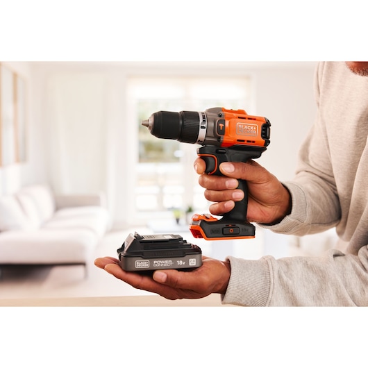 BLACK+DECKER BCD383D1 cordless drill and 18V battery held in hands, shown in a modern living room.