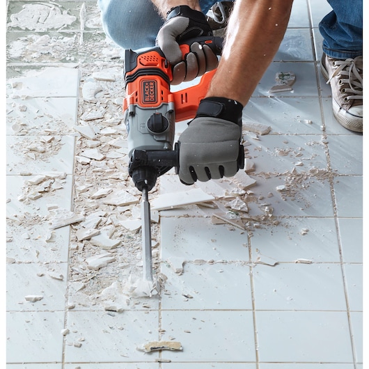 BLACK+DECKER BCD900B rotary hammer used to chisel tiles on a floor, close-up with gloves and debris.