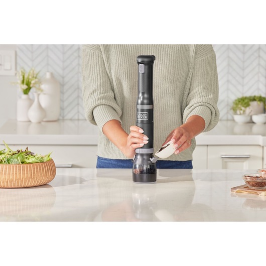 BLACK+DECKER BCKM1012KB electric grinder in black used in a kitchen, person pouring peppercorns into the device.