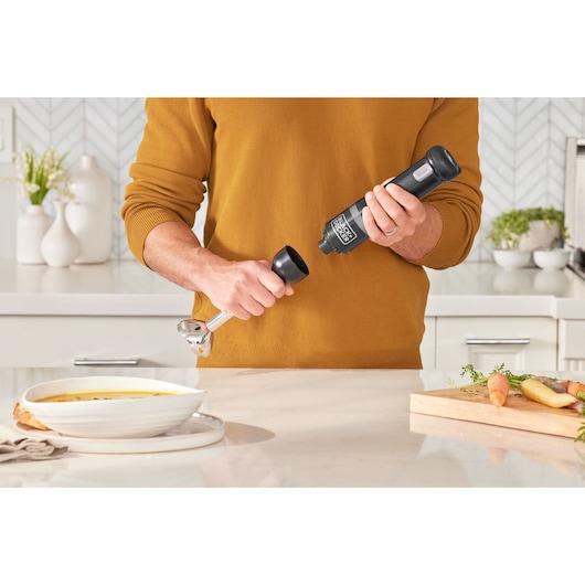 Photo of a person assembling a BLACK+DECKER BCKM1016KSB hand blender in a kitchen with a bowl of soup and vegetables.