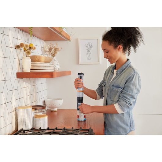Photo of person using BLACKDECKER BCKM1016KSG handheld mixer in a modern kitchen near stove and wooden shelves.