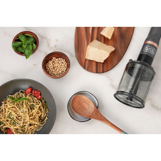 Photo of BLACK+DECKER BCKM101FP food processor with cheese, herbs, pine nuts, pasta, and wooden spoon on a kitchen counter.