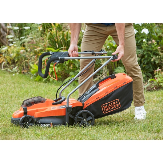 BLACK+DECKER BCMW3318L2 lawn mower in use on grass, orange and black design, manual handle adjustment.