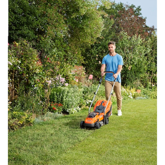 Photo of person using BLACK+DECKER BCMW3318N orange lawn mower on grass in a garden with lush plants.