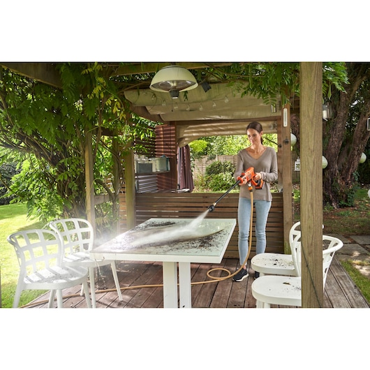 A person is using a BLACK+DECKER pressure washer to clean a dirty outdoor table on a wooden patio. The setting is in a garden with greenery and white outdoor chairs.