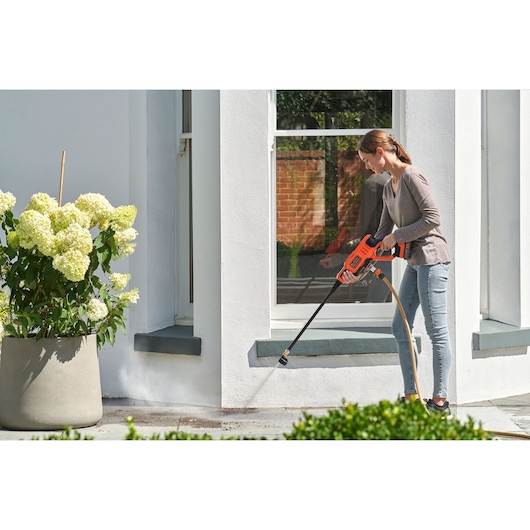 A person uses a BLACK+DECKER cordless pressure washer to clean an outdoor area near a white building, next to a large potted plant with white flowers. The pressure washer is orange and powered by a battery.