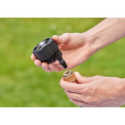 Close-up of hands holding two parts of a BLACK+DECKER device, likely a connector and nozzle, with a green blurred outdoor background.