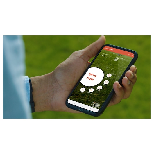 Photo of a hand holding a smartphone showing the BLACK+DECKER BCRMW123 robotic mower app screen, outdoors.