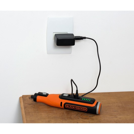 BLACK+DECKER BCRT8IK orange cordless rotary tool charging on a wooden table.