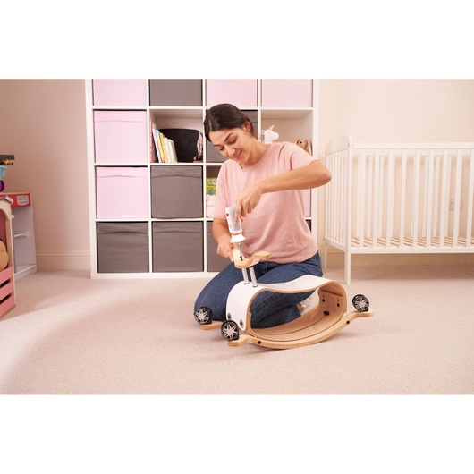 A person is assembling a wooden children's ride-on toy using a BLACK+DECKER tool in a nursery room with pink and gray storage cubes and a white crib in the background.