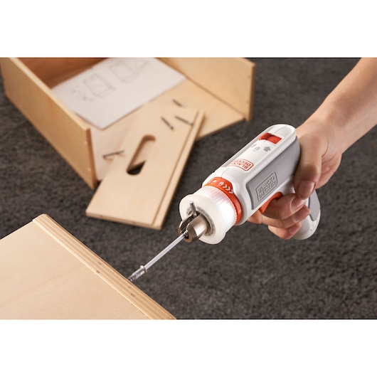 A person using a BLACK+DECKER electric screwdriver to assemble a wooden piece, with assembly instructions and hardware visible in the background.