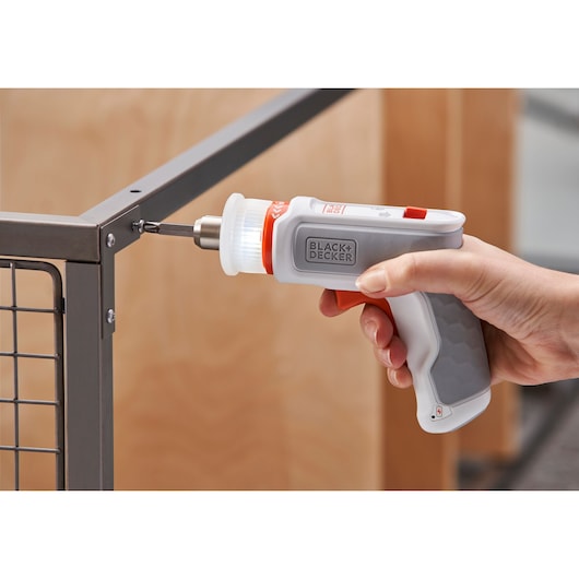 A hand holding a BLACK+DECKER electric screwdriver, using it to fasten a screw into a metal frame with a wire mesh.