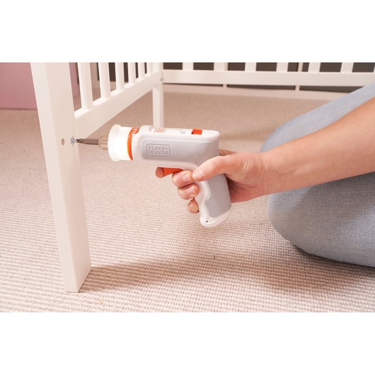 A person uses a BLACK+DECKER cordless screwdriver to assemble a white piece of furniture on a carpeted floor.