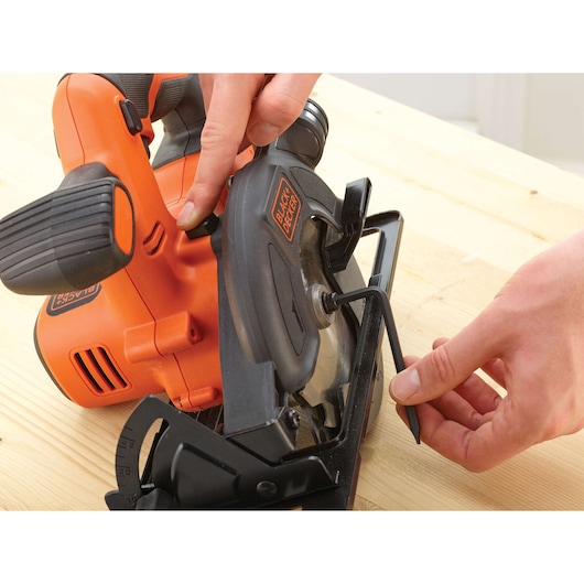 BLACK+DECKER BDCCS18 cordless circular saw close-up, person adjusting blade with hex key on wooden surface.