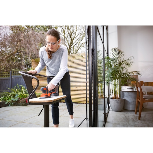 A person is sanding a wooden chair outdoors using a BLACK+DECKER BDCDS18 cordless sander. The chair is placed near a sliding glass door with a view of a modern interior and plants.