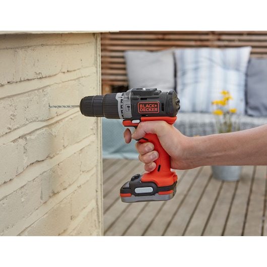 BLACK+DECKER BDCHD12S1 cordless drill in use on brick wall, orange and black design, outdoor setting.