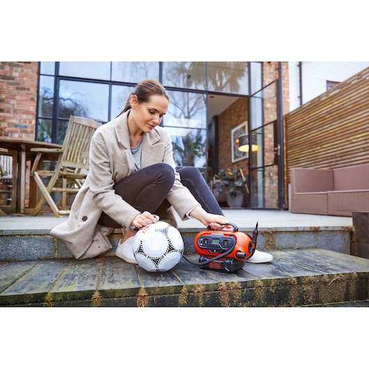 A person sitting outside on steps uses a BLACK+DECKER BDCINF18N air inflator to inflate a soccer ball. The portable orange and black inflator is placed next to the ball.