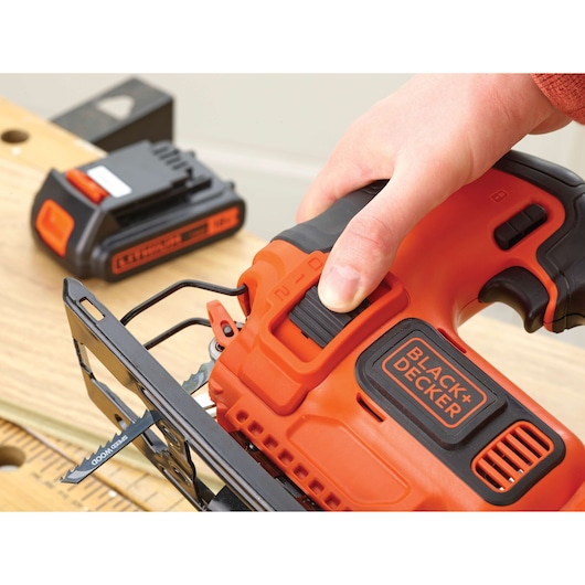 Photo of BLACK+DECKER BDCJS18M1 cordless jigsaw in use, close-up of hand on orange and black tool with battery pack.