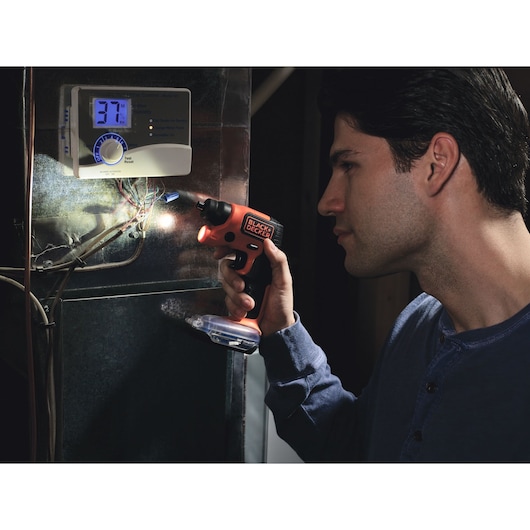 A person using a BLACK+DECKER cordless screwdriver to work on electrical wiring near an electronic thermostat or control panel.