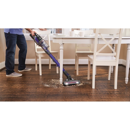 Photo of BLACKDECKER BDPSE1815P cordless stick vacuum cleaning debris from wooden floor near dining table.
