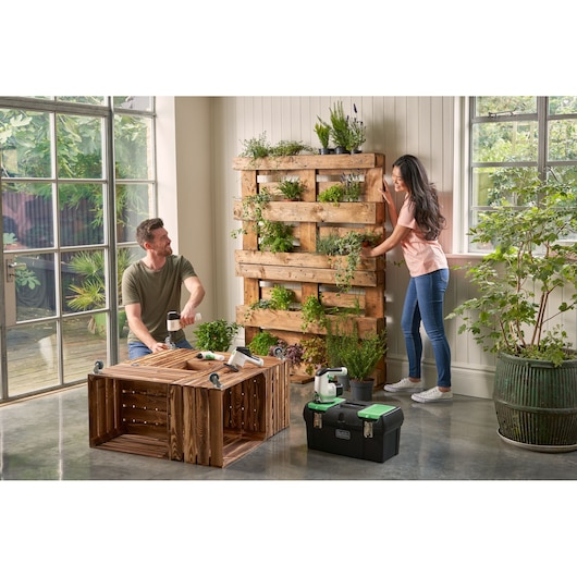 BLACKDECKER BDST60120 tool box on floor near two people building a wooden planter shelf in a bright indoor space.