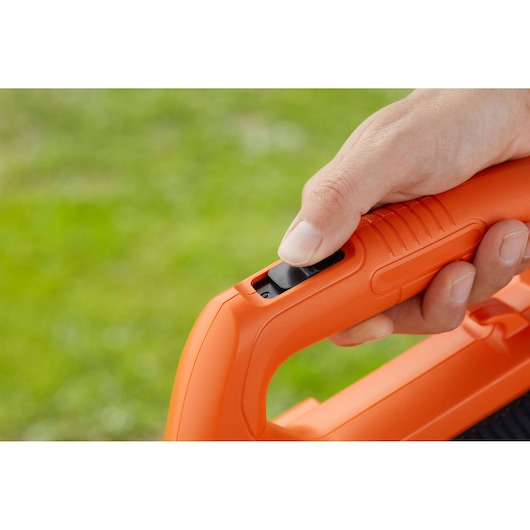 Close-up of a hand pressing the trigger on an orange BLACK+DECKER BEBL185 blower handle outdoors.