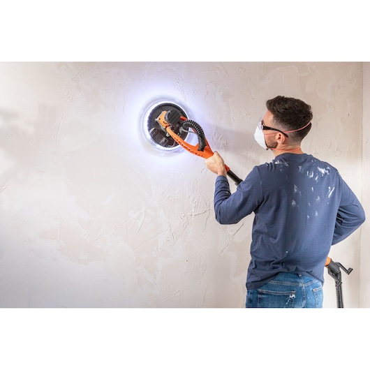 Person uses BLACK+DECKER wall sander with LED light on a white wall, wearing safety glasses and mask.