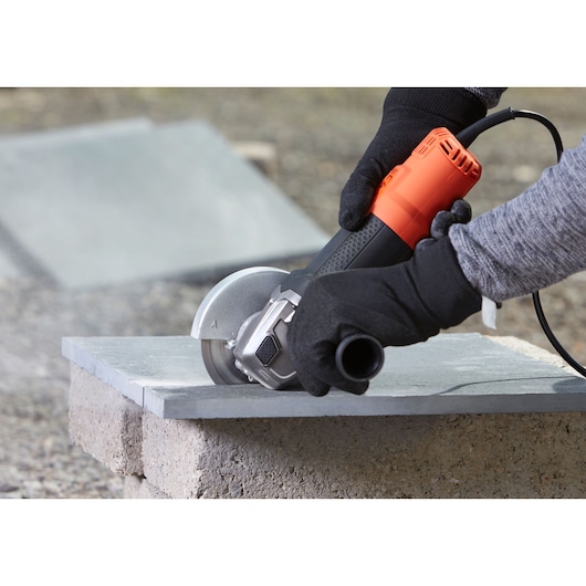 BLACK+DECKER BEG220K angle grinder being used to cut a stone tile, close-up with gloved hands.