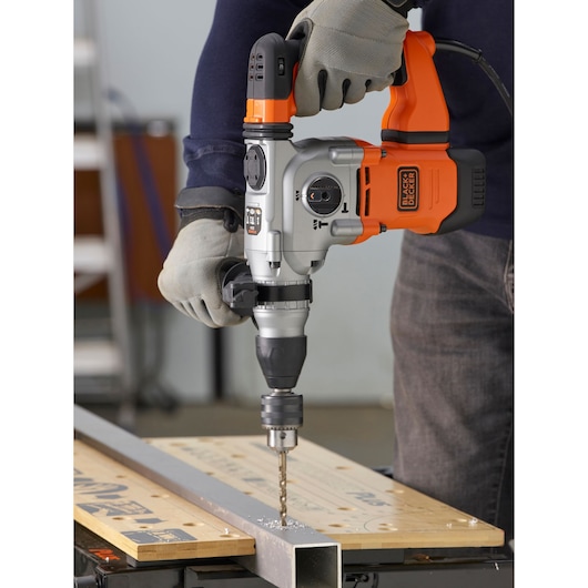 BLACKDECKER BEHS03K hammer drill in use, drilling metal on a workbench, with user wearing gloves.