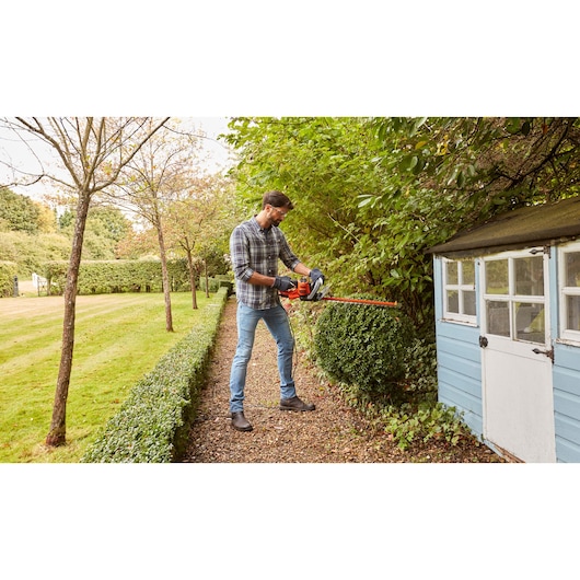 Person using BLACK+DECKER BEHTS455 hedge trimmer in a garden, trimming bushes near a small shed.