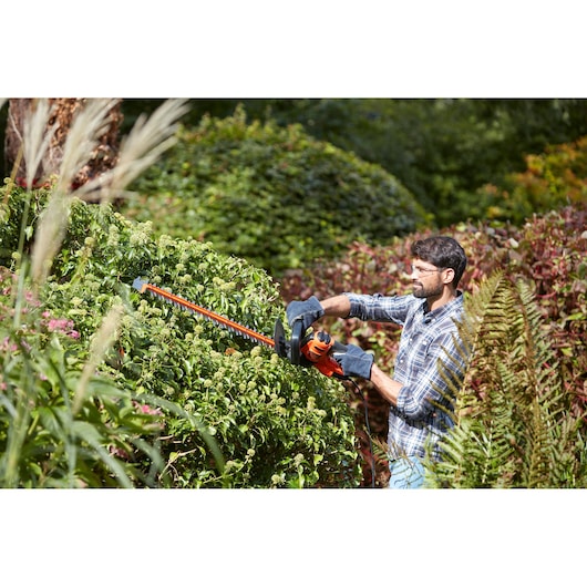 BLACKDECKER BEHTS501 hedge trimmer used outdoors with gloved hands trimming green bushes.