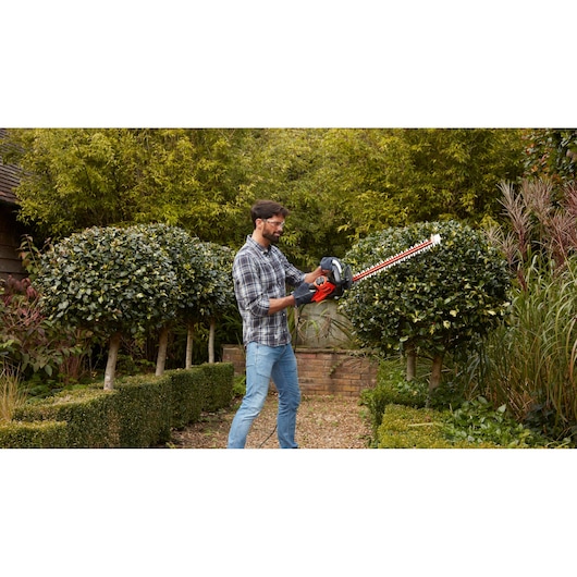 BLACKDECKER BEHTS501 hedge trimmer in use outdoors trimming bushes, red and black design visible.