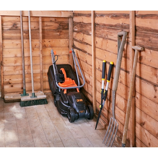 BLACKDECKER BEMW351 lawn mower with orange handle stored in wooden shed, surrounded by garden tools.