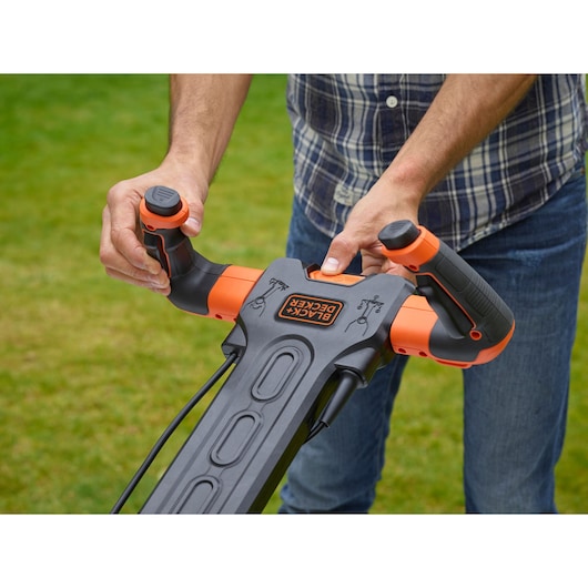 Close-up of person holding BLACK+DECKER BEMW461ES lawn mower handles with orange controls on grass.