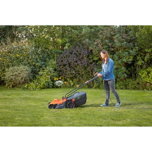 Person using BLACKDECKER BEMW461ES electric lawn mower with orange and black design on grass in garden.