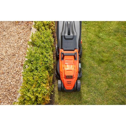 BLACKDECKER BEMW461ES orange lawn mower top view on grass beside hedge and gravel path.