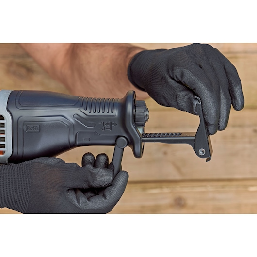 Photo of BLACK+DECKER BES301K reciprocating saw close-up, gloved hands adjusting blade clamp, wooden background.