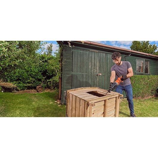 A person uses a BLACK+DECKER reciprocating saw (model BES301) to cut a wooden crate outdoors near a green shed. The individual is wearing gloves and working on the lawn.