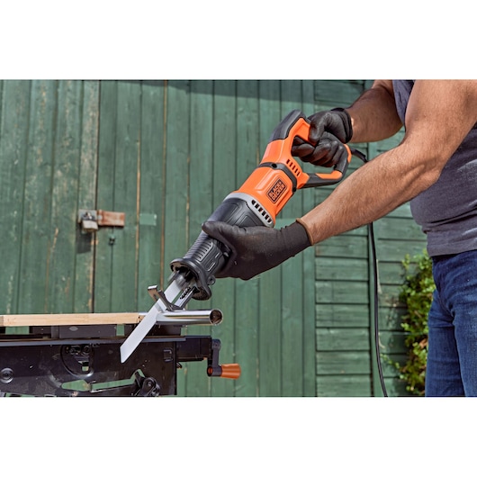 A person wearing gloves is using a BLACK+DECKER BES301 reciprocating saw to cut through a metal pipe, with the saw positioned on a workbench outdoors.