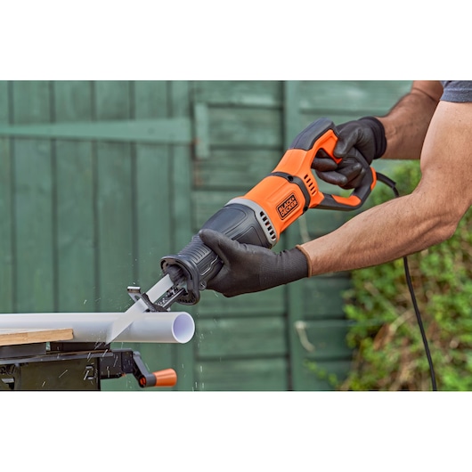 A person wearing black gloves uses a BLACK+DECKER reciprocating saw to cut a white plastic pipe outdoors.