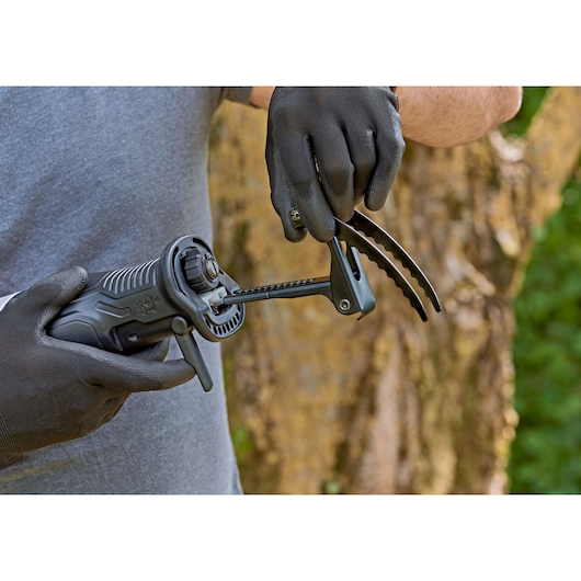 A person wearing black gloves is holding a BLACK+DECKER reciprocating saw with an accessory attachment outdoors, with a blurred tree trunk in the background.
