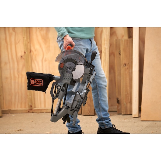 Photo of person carrying BLACK+DECKER BES701 miter saw with dust bag in workshop setting.