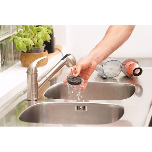 Person rinsing BLACKDECKER BHFEA18D1 vacuum filter under faucet with vacuum parts in stainless steel sink.
