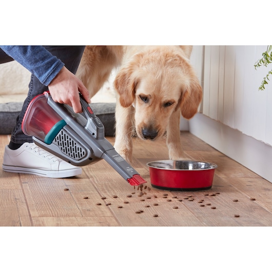 BLACKDECKER BHHV315J handheld vacuum cleaning pet food near red bowl on wood floor, side angle.
