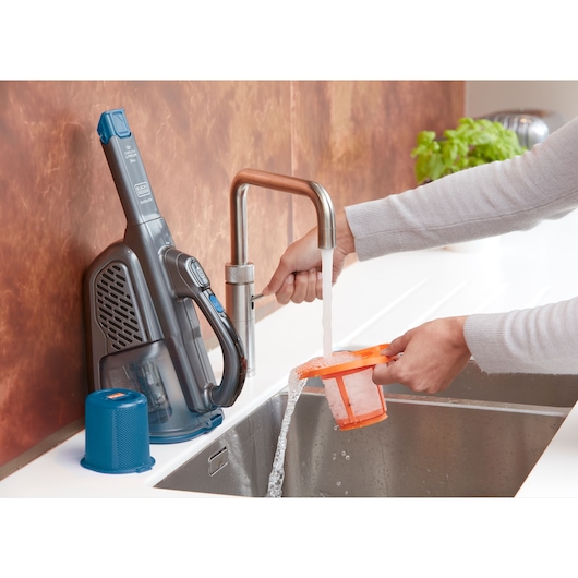 BLACKDECKER BHHV320J handheld vacuum filter being washed under kitchen tap, vacuum and filter parts on counter.
