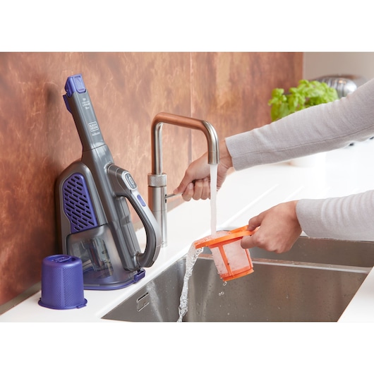 BLACKDECKER BHHV520BFP handheld vacuum cleaner filter being washed under a kitchen faucet, product and parts visible.