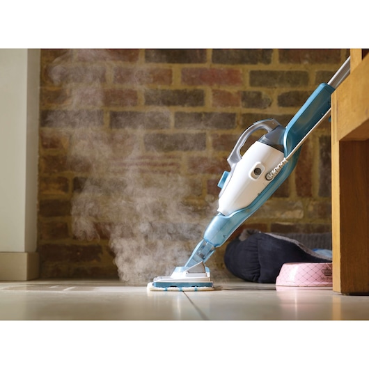 A steam mop with a blue and white design cleaning a tiled floor, releasing visible steam. The mop is close to a pet bed and bowl, with a brick wall in the background.